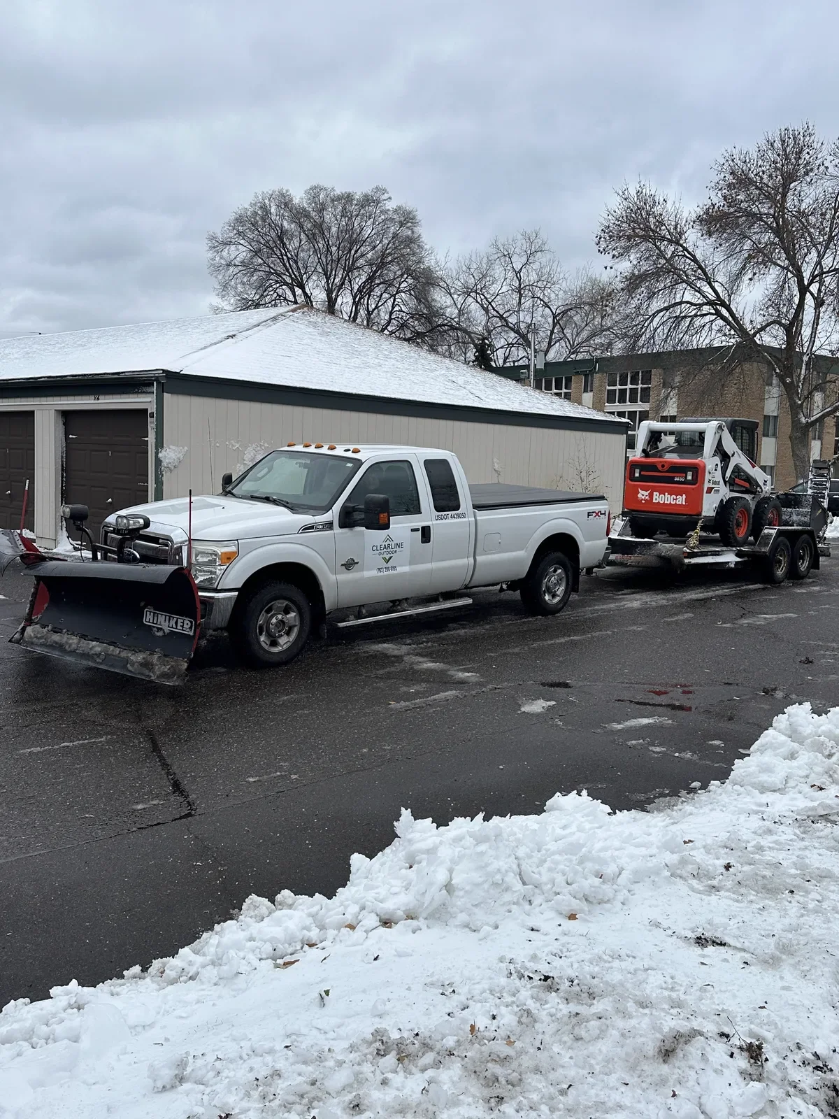 Clearline Outdoor truck with snow plow hauling a Bobcat skid steer, staged ahead of the North Twin Cities Metro winter season