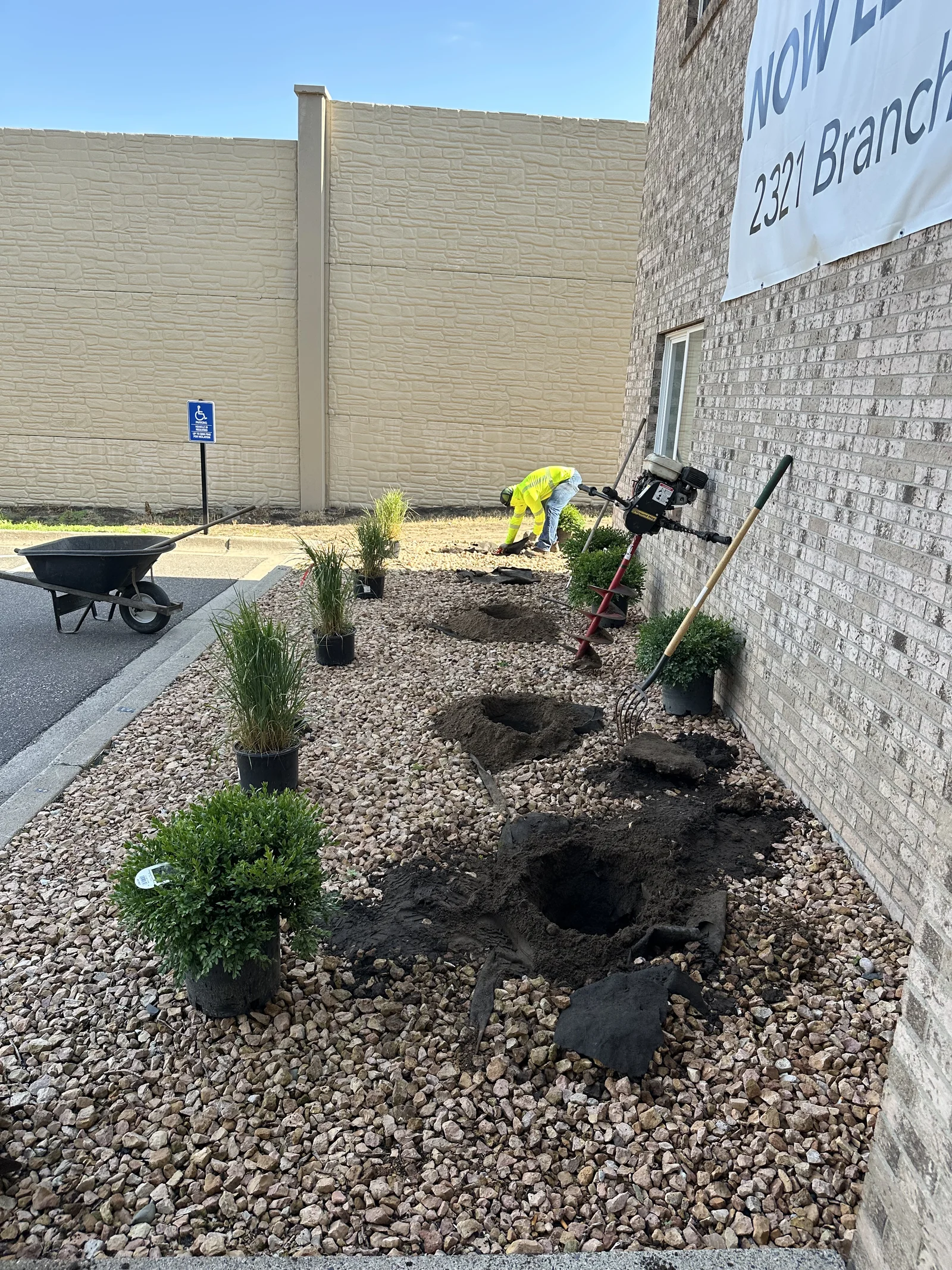 Crew member augering planting holes in a rock bed near a Now Leasing banner at Cove Apartments